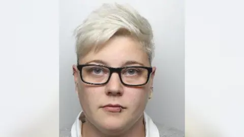 A mugshot of Sked who has short, dyed blonde hair and is wearing black-rimmed glasses. She has a piercing on the left side of her lower lip and a helix piercing.