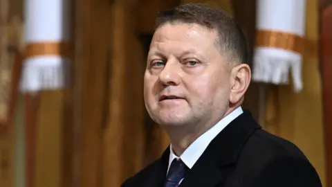 Valerii Zaluzhnyi seen wearing a black suit, white shirt and blue and red tie.