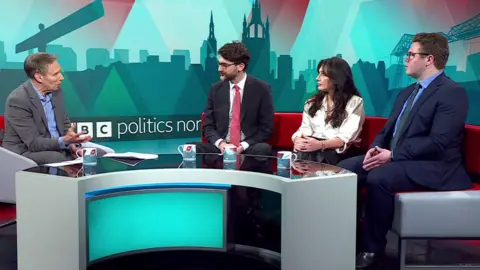 BBC Politics North presenter Richard Moss sits across from the red sofa that accomodates Markus Campbell-Savours, Jemma Joy and Anthony Mullen. The teal backdrop features silhouettes of regional landmarks. Moss looks quizzically towards Campbell-Savours while the other politicians watch on. 