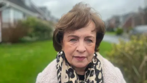 Dodds is looking at the camera. She has short curly brown hair. She is wearing a cream and black patterned scraf and a white coat.