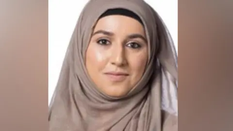 Birmingham City Council A woman smiling in front of a white background. She is wearing a mink coloured hijab covering her hair and shoulders.