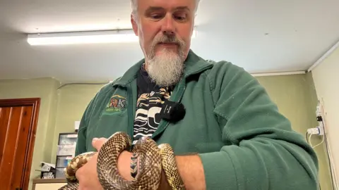 Jamie Mintram has a grey beard and grey hair, he's looking down at his hand. Micky the snake has wrapped himself around Jamie's hand. Jamie is wearing a green fleece jumper.