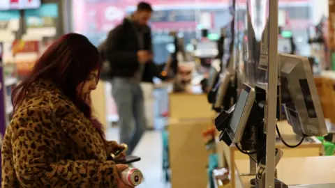 Andy Rain/EPA-EFE/REX/Shutterstock A woman in a patterned coat is standing at a self-checkout in a shop, holding a can of drink.