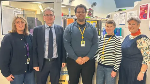 Team of 4 volunteers and the Police Crime Commissioner at Perry Barr Custody Suite. A woman on the far right has blonde hair and is wearing a navy blue jacket and blue jeans. She's stood next to the PCC who is a man and is wearing a black suite, with a blue shirt and black tie. Stood to his right, and in the middle, is a man with a blue jumper on and black trousers. Next to him is a woman wearing a black and white striped jumper and blue jeans. And next to her is a woman with short grey hair, orange scarf, floral shirt under a denim jumpsuit.