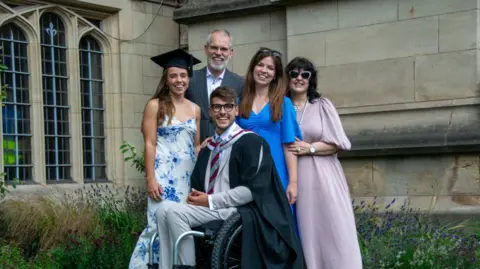 PA Media/University of Bristol Xander in his graduation gown, sitting in a wheelchair, with his wife and three family members around him, smiling at the camera, outside a building at the University of Bristol