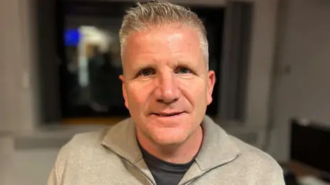 A man with short white hair looks into the camera. The background is blurred. The man is smiling and is wearing a light grey zip jumper with a dark tshirt.