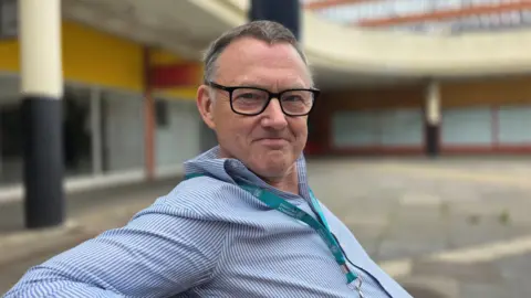 Paul Moseley/BBC Mike Stonard is sitting on a bench in Anglia Square. He's wearing a blue and white pinstripe shirt along with a light blue lanyard. He also is wearing glasses.