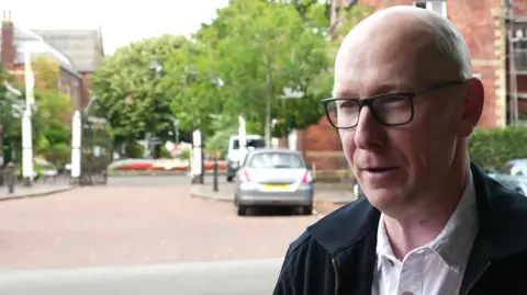 Southport MP Patrick Hurley with a bald head wearing black glasses, a white shirt and black jacket near the entrance to the Botanic Gardens in Churchtown, Southport.