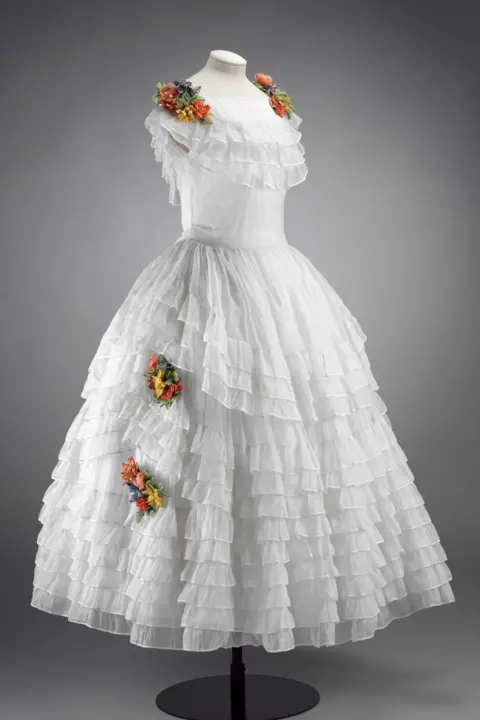 Sotheby's A white 18th-century-style dress with multiple tiers of sheer ruffles and clusters of colourful artificial flowers on the shoulders and skirt, displayed on a mannequin.