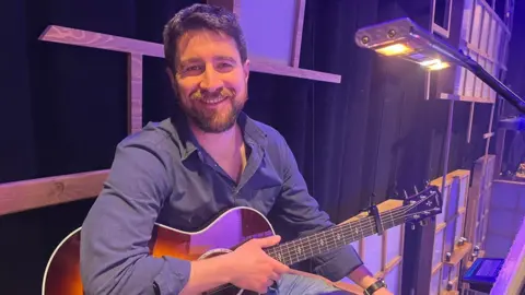 Robin Hiley is seen in a blue shirt and jeans sits on an elevated part of the wooden set, holding an acoustic guitar.