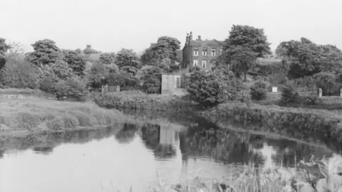 Bolton Archives A black and white photograph of Rock Hall. The image shows the manor house overlooking the River Croal, with reflections of trees in the water. 