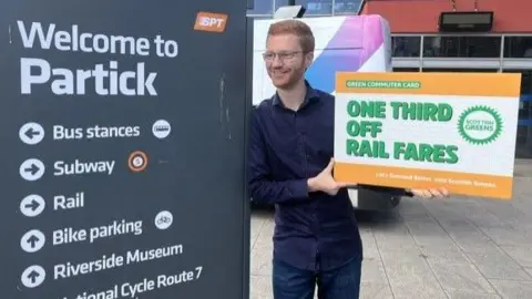 Man standing beside a “Welcome to Partick” transport sign, holding a large sign that reads “One third off rail fares,” outside a station entrance with a bus visible behind him