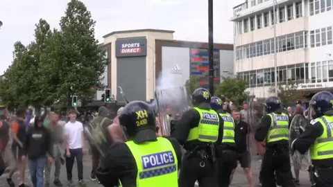 Police officers in riot gear face rioters during the unrest that followed the stabbing of three children in Southport last summer. The police officers are holding large clear shields, facing down would-be rioters. A Sports Direct store stands on a street corner in the background.