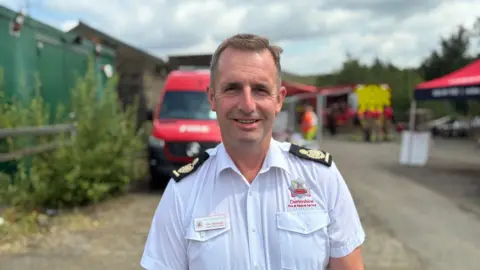 BBC/Mark Ansell Dept Ch Fire Officer, Clive Stanbrook with short brown hair and white uniform with fire vehicles behind him, bushes and a tent.