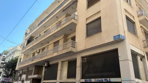 PA Media 9 Rethymnou Street in Athens, where Elisabeth-Jane Ross was staying before she was killed - Exterior view of a multi-storey beige apartment and commercial building on a street corner. The building features several balconies with metal railings, shuttered windows, and air-conditioning units. At street level, large dark shopfront windows display faded signage and graffiti. A blue street sign with Greek text is mounted on the corner of the building, and the scene is lit by bright daylight under a clear blue sky.