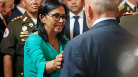 Delcy Rodriguez is wearing a green suit and glasses while greeting greets aa man whose back is toward the camera. sShe has her hand on his arm and is smiling. A man in uniform and a crow is behind her