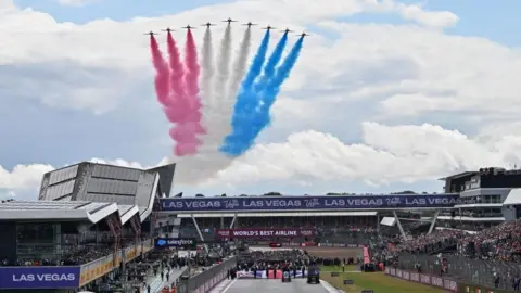 EPA Nine red arrows with red, white and blue smoke behind them fly over stadiums at the Silverstone Circuit. The circuit is full of fans.