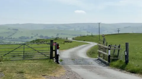 A narrow country lane weaving through a grassy hillside, with a wire fence and a gate