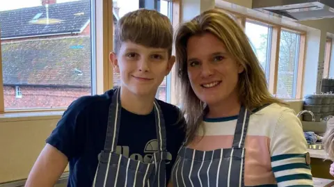 Photograph of Ellen Roome and her son Jools. They are stood in a kitchen area with blue and white stripped aprons on.