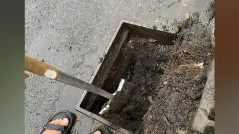 Julie Warne A spade digging inside a road drain which is full of mud and debris.