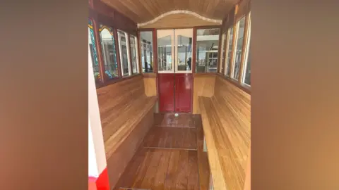 David McKay The interior of the wooden carriage. It has an arched roof and long in-built wooden benches on either side. On the far end, there is a red and white door and there are window panels all the way around.