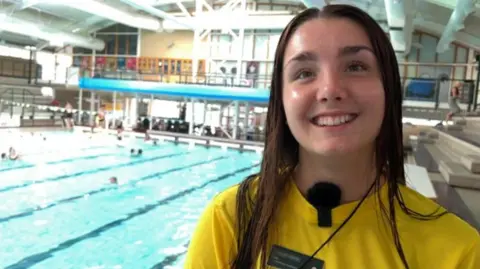 Lilly. She is pictured in front of a swimming pool. She has long dark hair and is smiling at the camera. She is wearing a yellow t-shirt