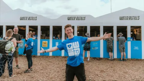 WaterAid/ Lucy Le Brocq Conor Blundell wearing a blue wateraid t-shirt, standing with his arms outstretched and smiling in front of a water refills station at Glastonbury