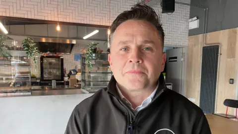 Brian Reid. He has dark hair and blue eyes, wearing a black zip up jacket with white shirt underneath. A cafe counter is behind him.