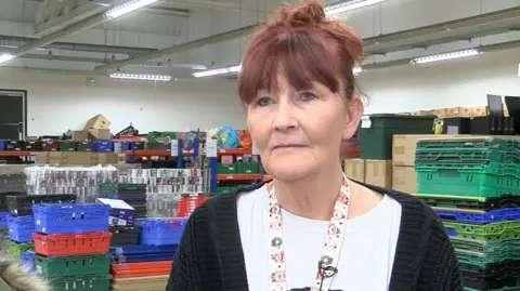 Andrew Sinclair/BBC A woman wearing a white t-shirt with a snowman on it and a black cardigan on top stands in front of crates full off donated food. 