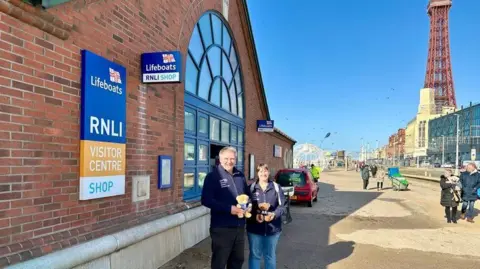 Allan Thornhill Allan and Helen Thornhill standing outside the RNL lifeboat station in Blackpool with the seafront promenade and Blackpool tower behind them 