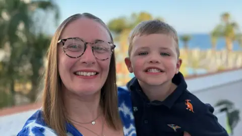 Jennifer Taylor, holding her son William. Jennifer is wearing a blue-and-white floral dress and has long brown hair. She is wearing tortoiseshell glasses. William is in a navy shirt with dinosaurs embroidered on it.