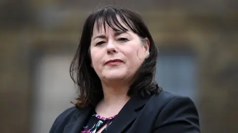 Michelle Gildernew looking at the camera with a serious expression and her head tilted up slightly.  She has shoulder-length dark hair with a fringe and is wearing a black, purple and green top under a black suit jacket.   The background is blurred. 