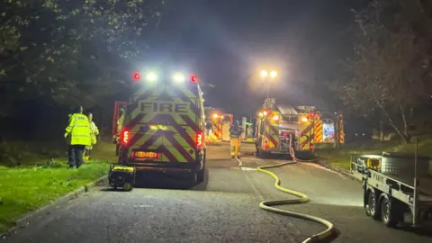 Pete Hamilton Multiple fire engines and vans parked along a dark road. There is a yellow hose running down the centre of the road. There is a bright, tall light in the distance and two people in yellow hi-vis stand on the grass verge on the left.