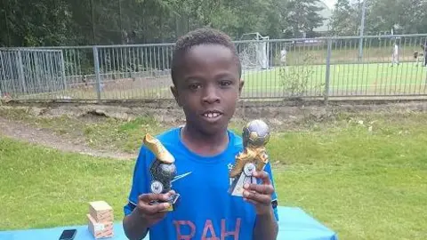 Othniel Adoma wearing football kit and holding two trophies.