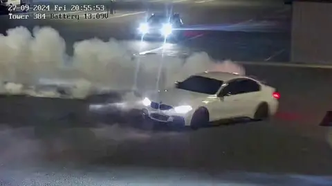 A white car turns sharply in front of a trail of exhaust smoke 