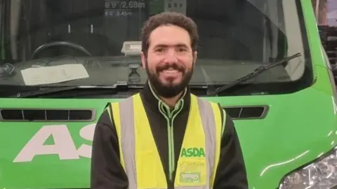 Asda A man with dark brown hair and beard, wearing a yellow high vis with ASDA written on it and stood in front of a green van. 