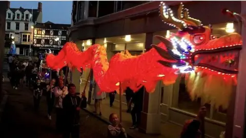 It is dusk and people are parade in the streets. An illuminated red dragon is being held aloft on poles by some of the people in the parade. 