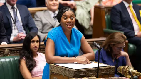 Kemi Badenoch in the House of Commons at the despatch box. She is wearing a blue dress and behind her are Conservative MPs.