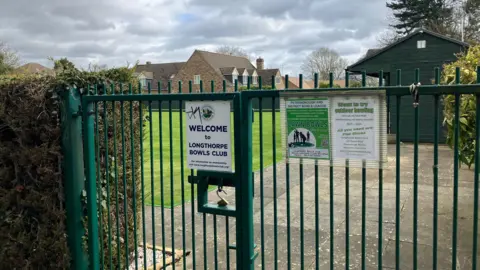 Shariqua Ahmed/BBC Entrance to the bowls club - a green gate with white sign boards on it.