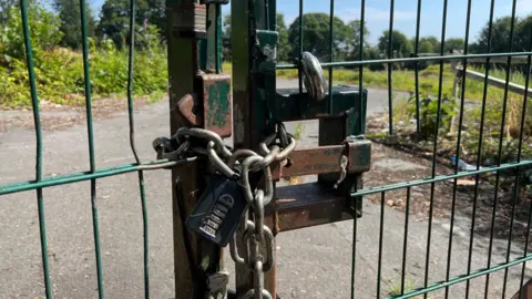 A large green wired gate, padlocked and chained. In the background is greenery and wasteland.
