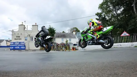 Classic TT Two racers on the exit to Ballaugh Bridge. The first is riding a black motorcycle while the second is on a green machine. Both have wheels off the ground. The Raven pub is in the background.