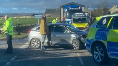 John Rhind A crash scene with a grey car in the centre badly damaged and a number of emergency vehicles surrounding it 