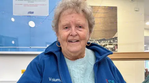 Sheila Underwood with short grey hair wearing a blue fleece, pale blue jumper and gold hoop earrings in a community centre. She is sitting at a desk with a noticeboard behind her. She is smiling.