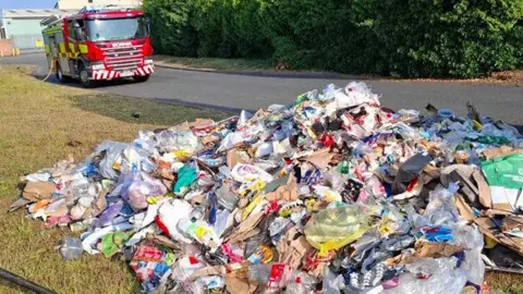 Herefordshire Council A large pile of rubbish on some grass next to a road. There is a red and yellow fire engine in the background.