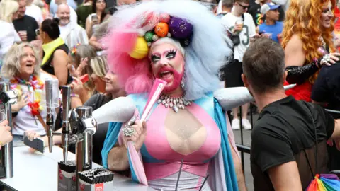 Stuart Rhoades A drag performer wearing a large multicoloured wig and a pink white, and blue lycra costume is pretending to sing into a fan. Crowds of people can be seen in the background. 
