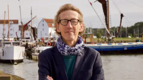 Contributed Nick Cottam smiles at the camera as he stands near the River Deben in Woodbridge. Boats are moored behind him. He has short light brown hair and wears a navy jacket, green jumper and black and white scarf, 