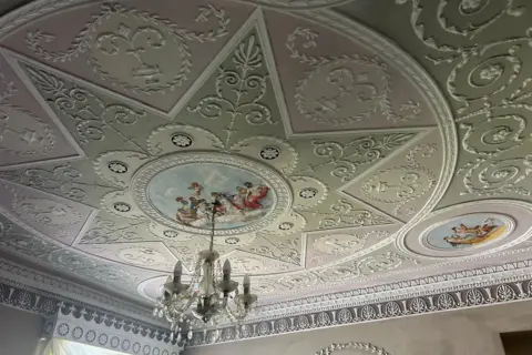 A close up of one of the ceilings inside Mr Jones' home. The ceiling is painted a variety of different pastel colours with intricate white neo-classical detailing. In the centre of the ceiling there is a circle depicting god-like figures sitting on a cloud, with the sky around them. A chandelier hangs from the circle. A large concentric circle is around the artwork, and within that is a smaller star shape, each containing ornate plasterwork lines and patterns.