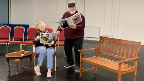 David and Jean Young rehearsing their comedy act. David is standing up, and reading a magazine, while Jean is sat down next to him, also reading a magazine.