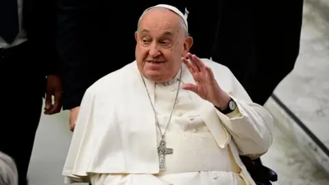 Pope Francis waves and smiles at onlookers as he is pushed in a wheelchair. 