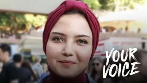 Susan Nasser, a young woman wearing a burgundy turban, pictured outside with people in the background. The words YOUR VOICE have been imposed on the photo on the right hand side.
